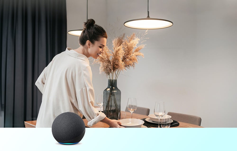 A woman arranging place settings at a table with a tray, next to a black Amazon Echo smart speaker on the tabletop