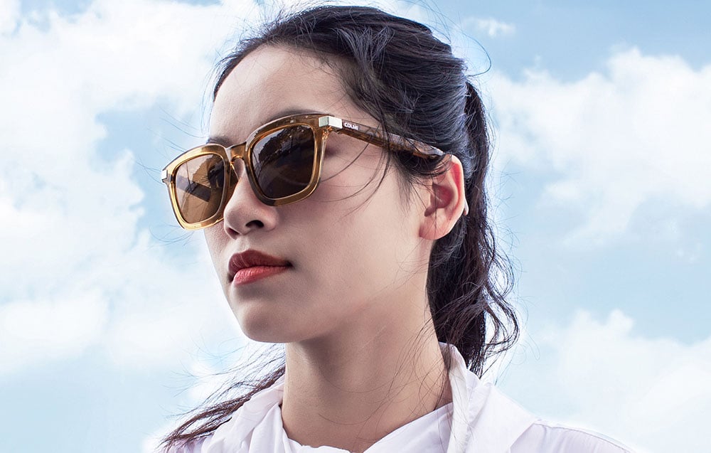 Woman in profile wearing COLMI smart glasses against a bright, cloudy sky.