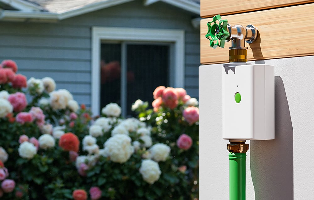 Smart irrigation controller mounted on a building wall, with colorful plants in the background