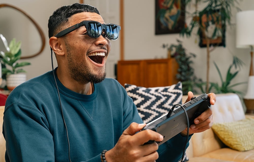 A man in XR glasses laughs while playing video games with a controller in his hands in the living room