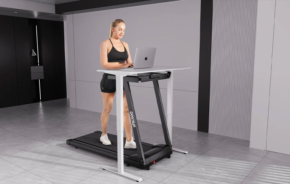 A woman walks on a treadmill next to a desk with a laptop in a minimalist room