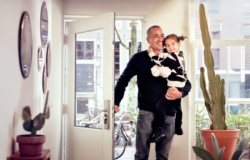 A smiling man holding a small child in his arms enters the house through an open white door