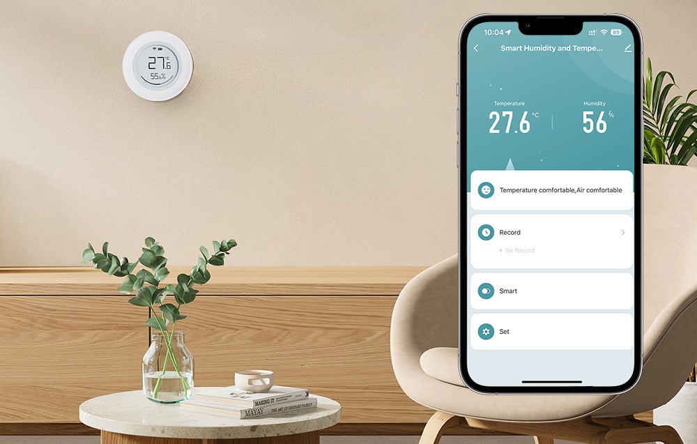 A sensor mounted on the wall above a dresser, next to a smartphone showing temperature readings of 27.6°C and humidity of 56%