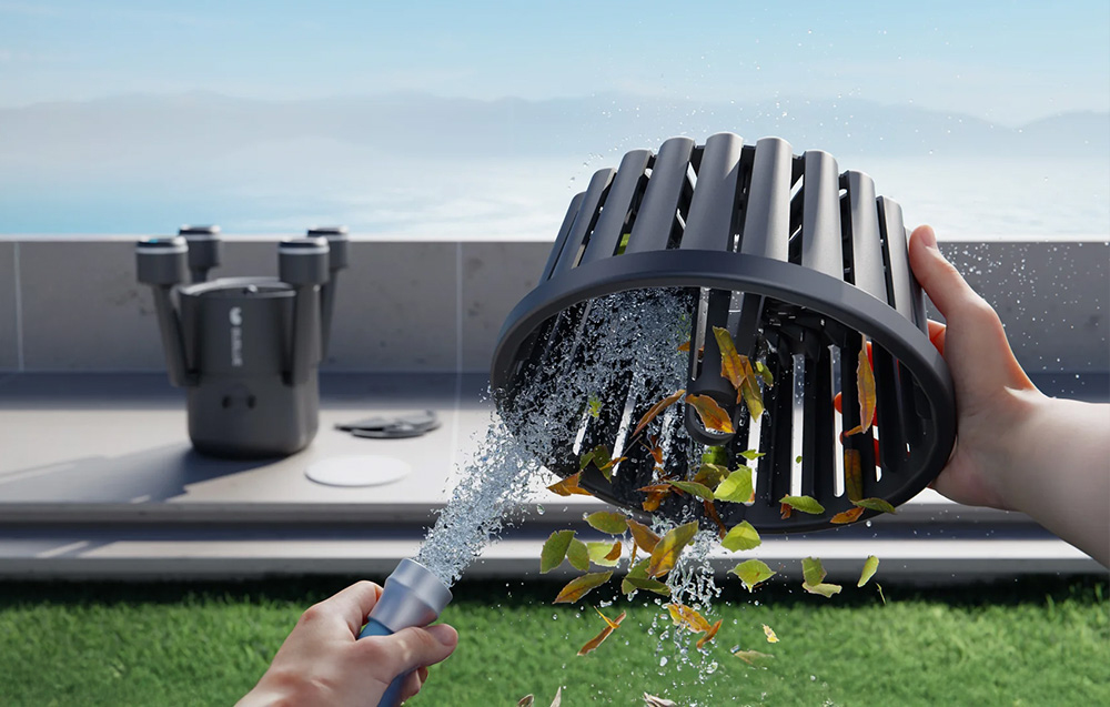 Hands hold the robot’s filter basket, rinsing it with a stream of water from a garden hose at the pool’s edge
