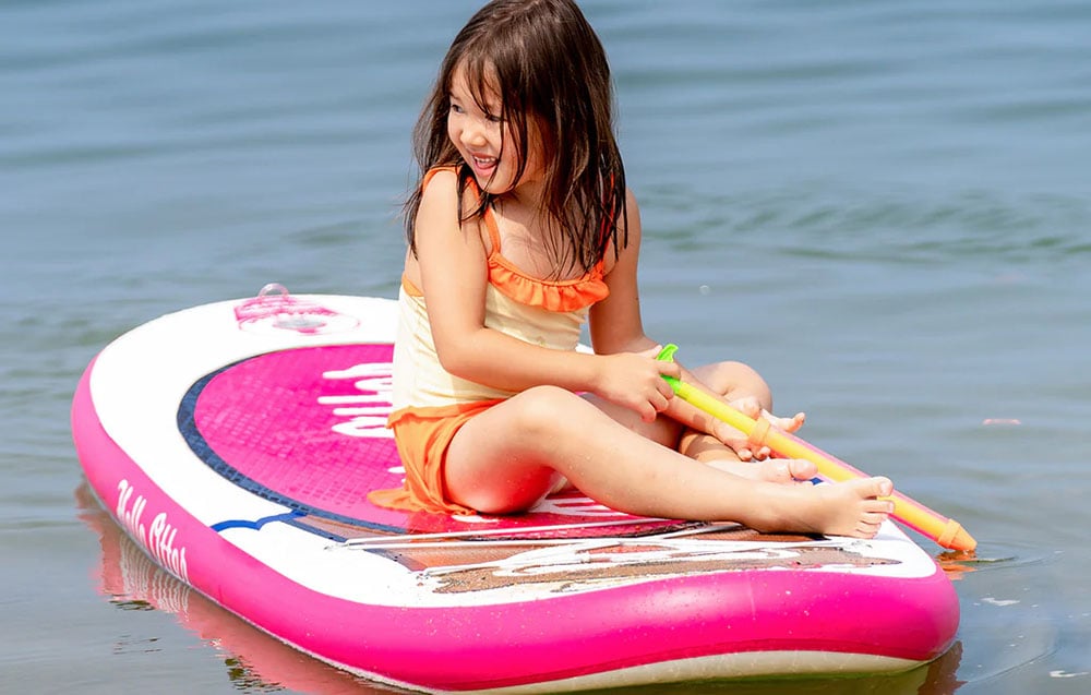 A girl with a water gun in her hand sits on a white and pink surfboard