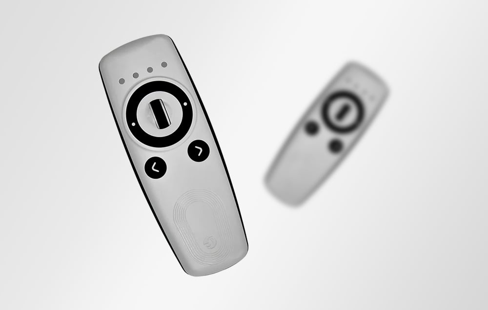 White Shelly remote control with black knob and two directional buttons on a light background, with a fuzzy duplicate in the background.