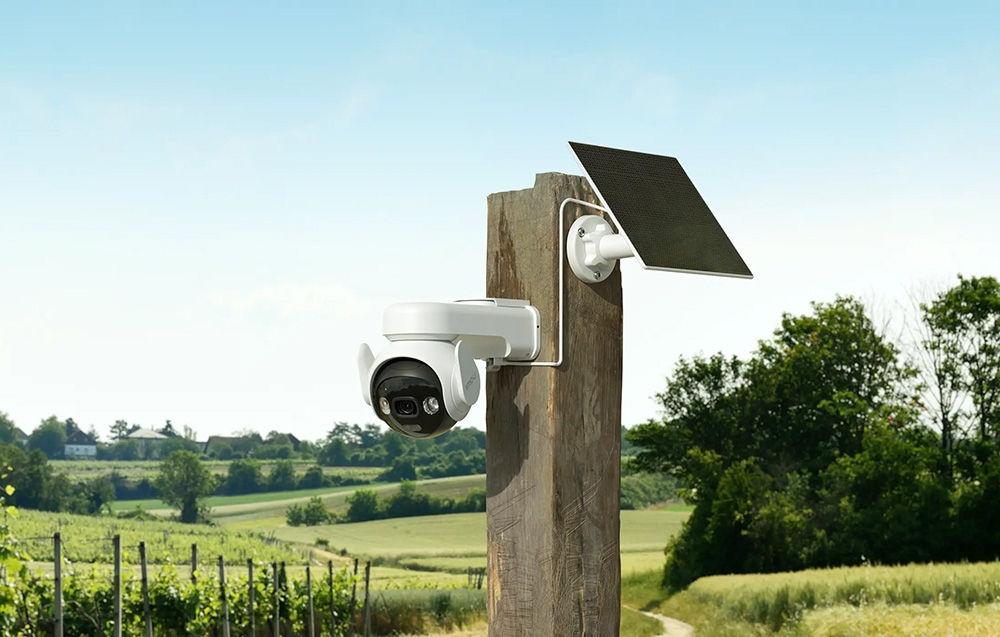 A white camera with a solar panel mounted on a wooden pole against a backdrop of fields, vineyards, and a blue sky
