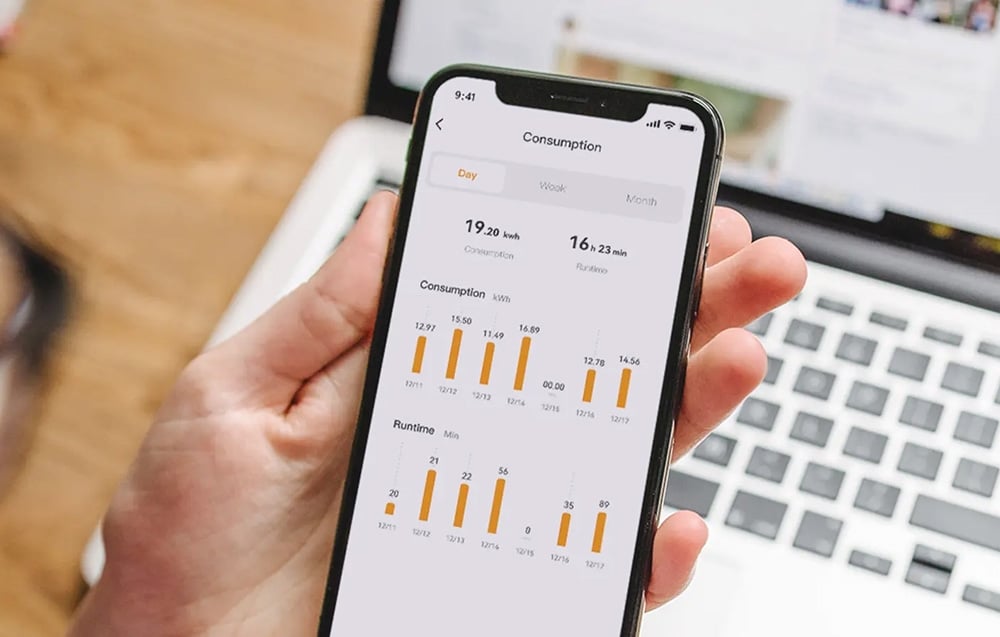 A hand holding a smartphone with the Consumption app displaying graphs of electricity consumption and operating time