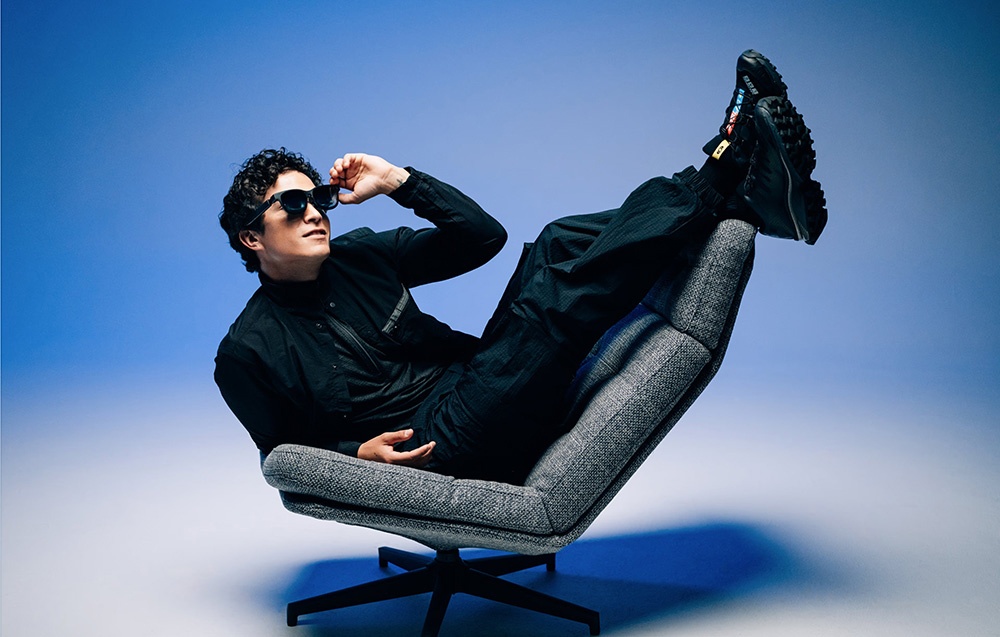 A man in black clothing and modern glasses lies relaxed on a gray armchair.