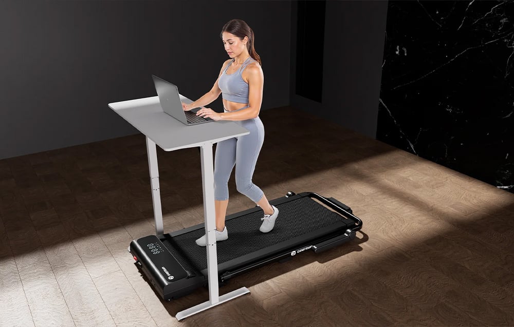 A person in a gray outfit works on a laptop at a white desk next to a black DeerRun treadmill