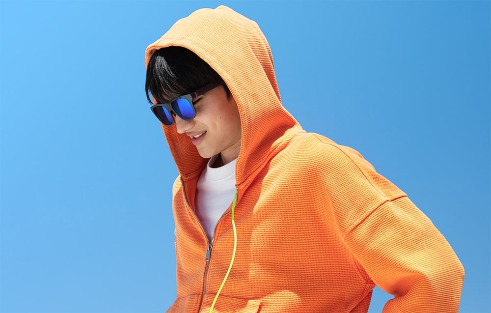 A man in an orange hoodie and glasses with blue lenses is looking down against a backdrop of a blue sky
