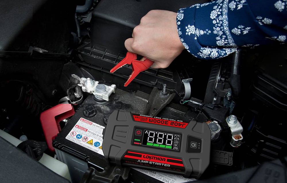A hand holding a red clamp over a car battery next to the jump starter lying on the ground.