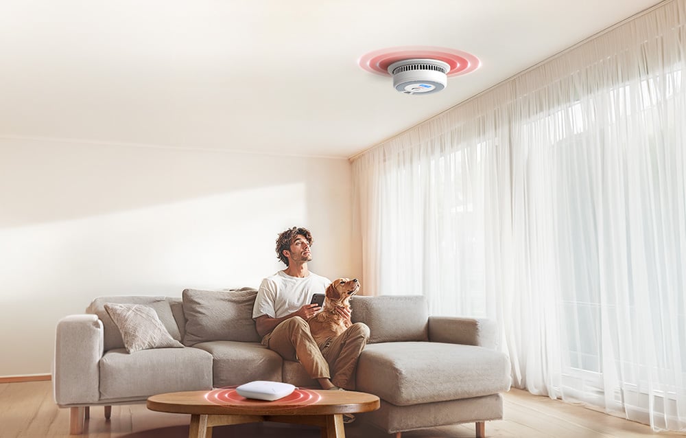 A man with a dog sits on the couch in the living room, looking at the smoke detector on the ceiling and holding a smartphone