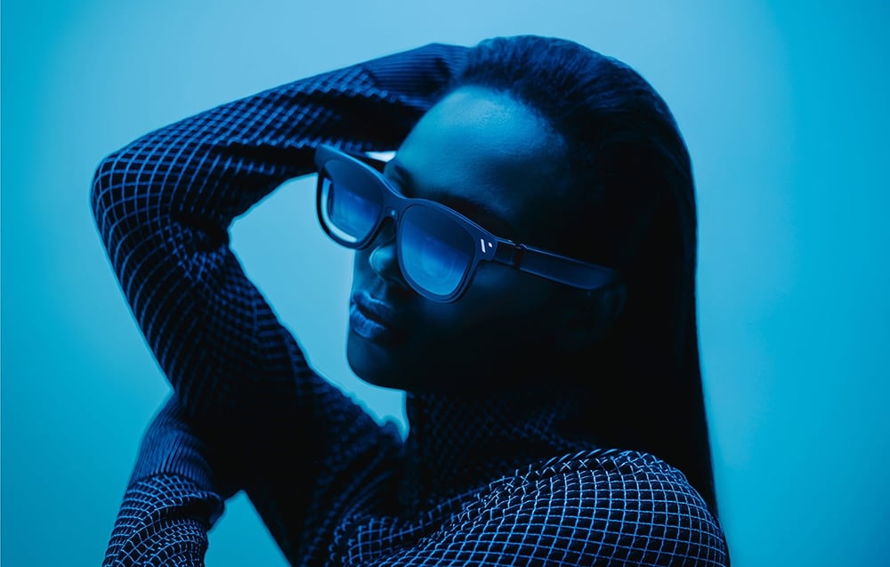 Woman in sunglasses with hand behind head, posing in blue studio light