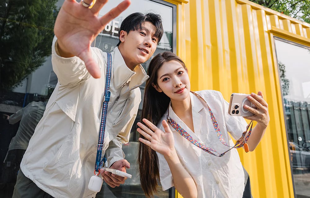 A man and a woman stand perched next to each other against a yellow building, wearing PGYTECH straps around their necks