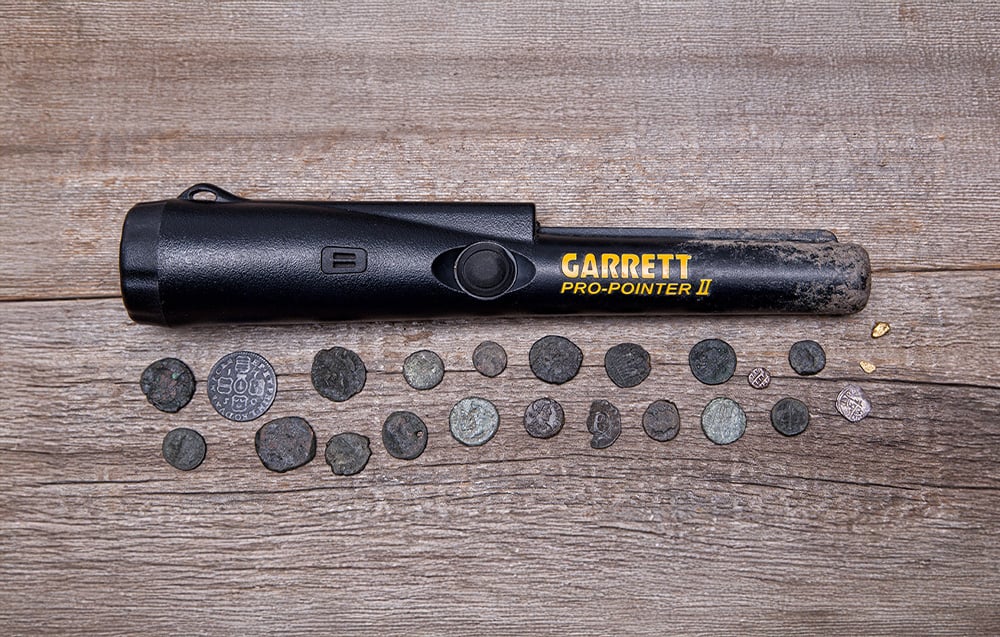 The metal detector lies on wood over a row of old coins and gold.