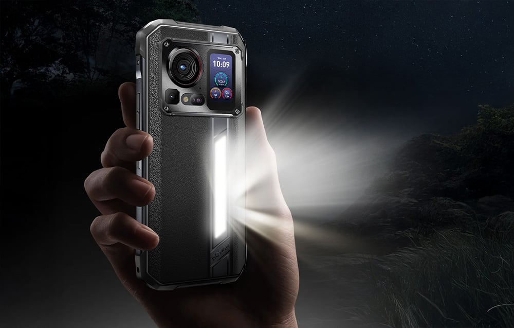 A hand holding a black smartphone with a powerful vertical LED flashlight on the back cover turned on in a night scene.