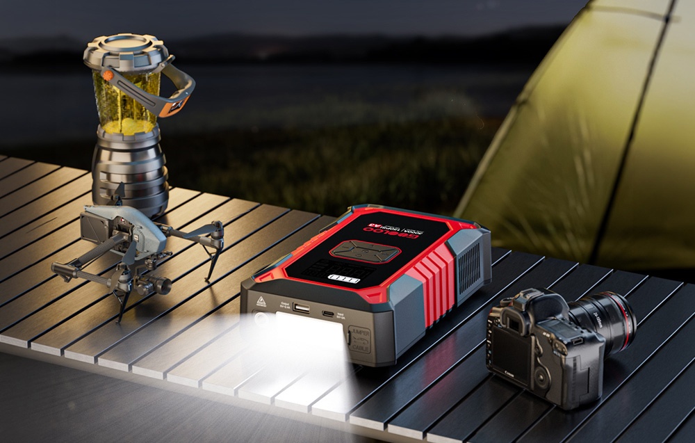 A jump starter with its flashlight turned on is lying on a table next to a drone, a camera, and a camping lamp