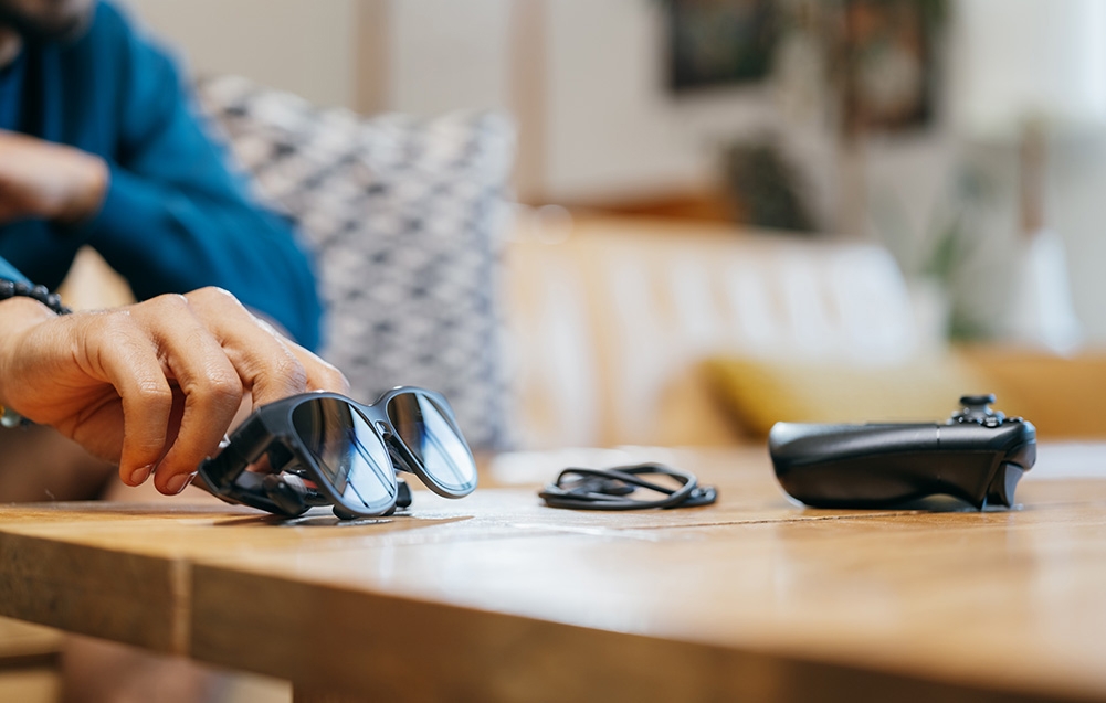 A hand reaches for black XR glasses lying on a wooden table next to a cable and a game controller