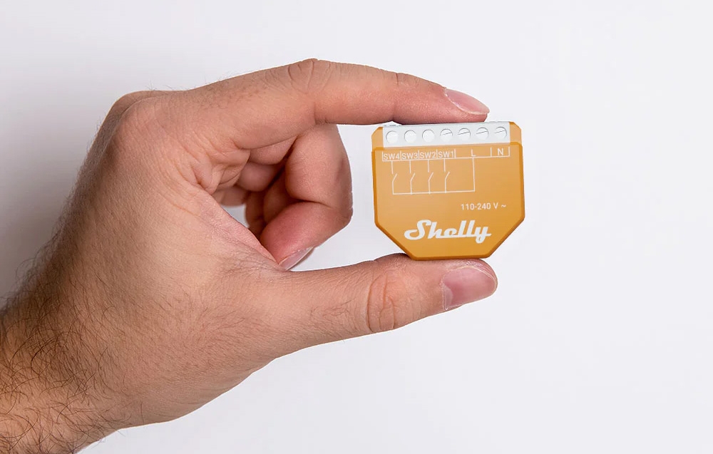 A hand holding an orange Shelly module with the Shelly logo and connectors against a white background