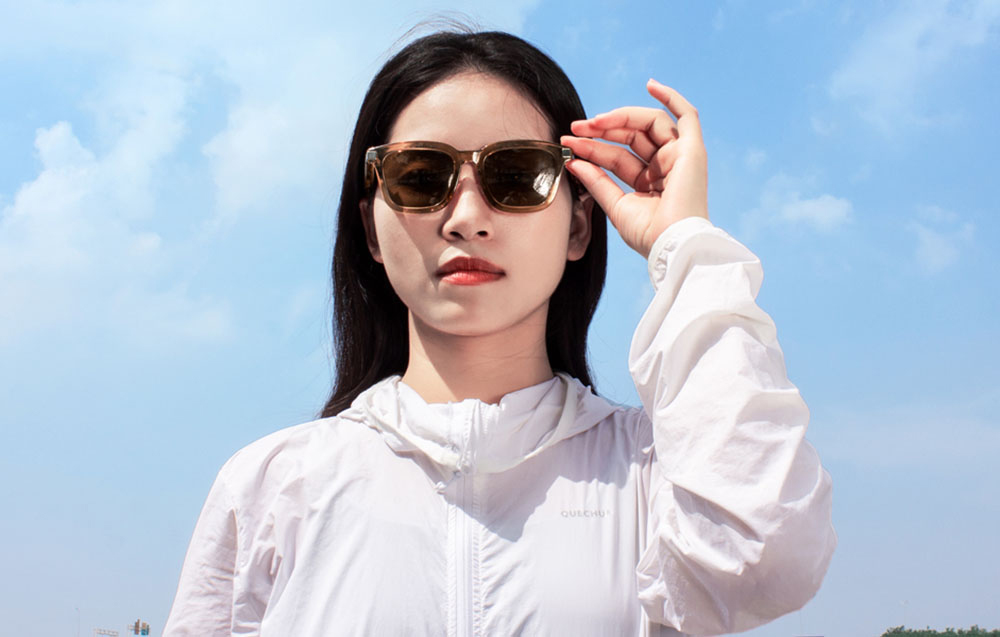 A woman in a white sports sweatshirt looking directly into the camera and holding her hand on the temple of her smart glasses.