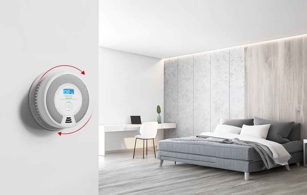 A carbon monoxide sensor mounted on the wall in a modern bedroom, with red arrows suggesting the installation