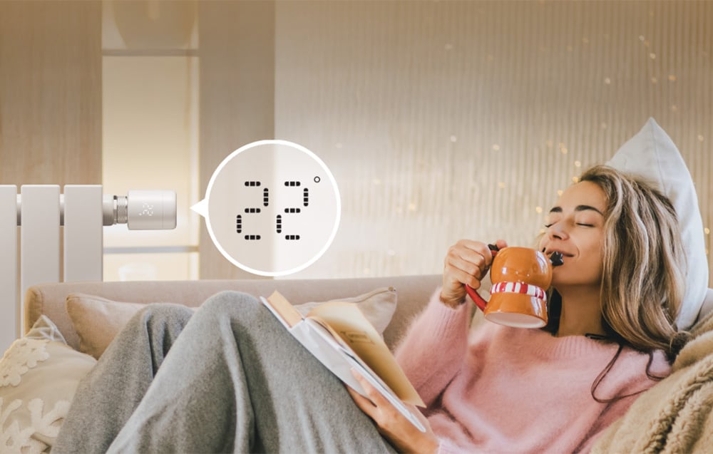 A woman lies on a couch with a cup in her hand, behind her a radiator with a temperature of 22 degrees Celsius is visible