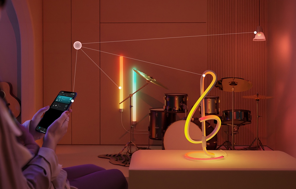 A hand holds a smartphone with an app, from which lines lead to the glowing lamp and a drum set in the background.