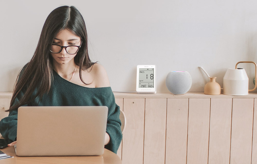 A woman wearing glasses is working on her laptop, behind her on the shelf is an air quality meter and a voice assistant