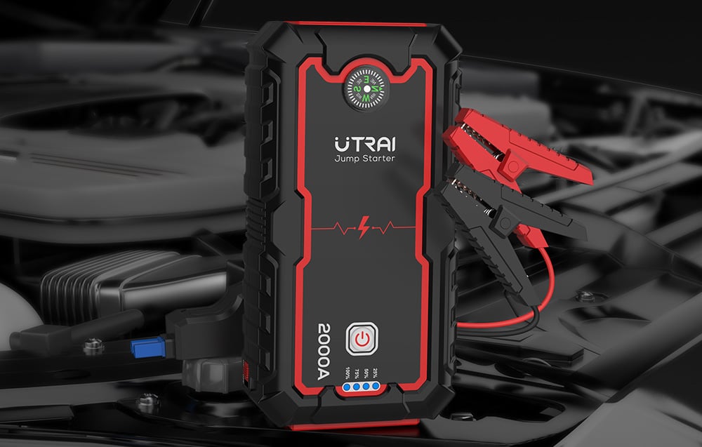 A black-and-red Utrai jump starter with connected cables and clamps, set against the background of a car engine.