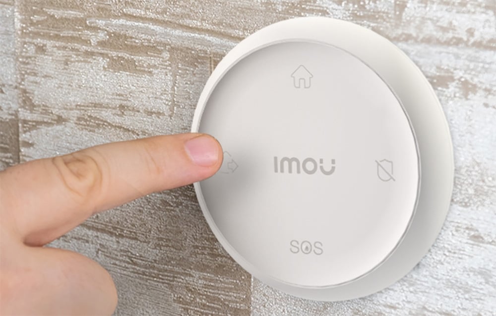 An index finger presses a button on a round white Imou control panel featuring icons of a house, a shield, and the word