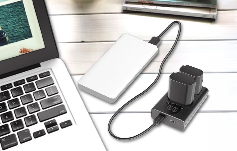 A dual-battery charger connected via a USB cable to a white power bank next to a laptop on a light-colored tabletop.