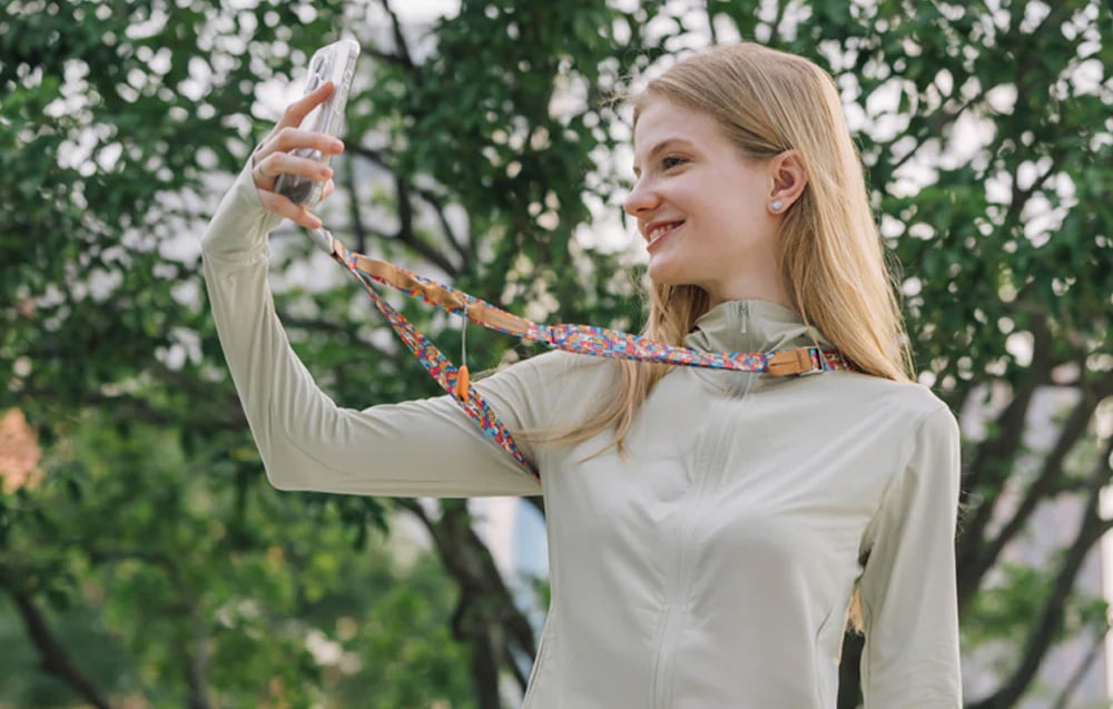 A young woman takes a selfie with a smartphone mounted to a PGYTECH strap that hangs around her neck