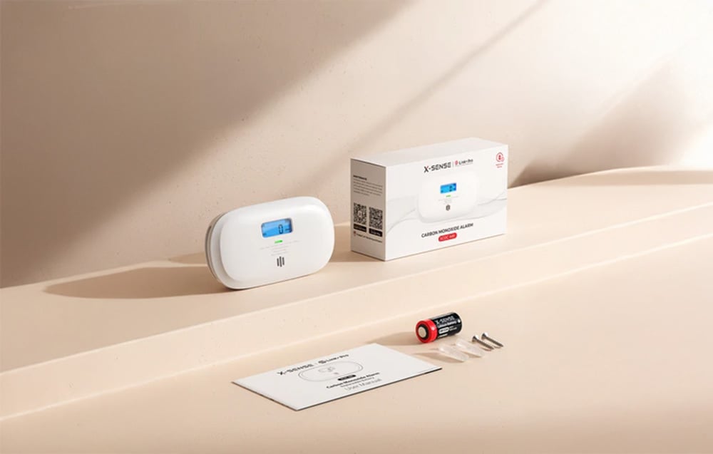 X-Sense carbon monoxide detector in white case with blue display next to box, instructions, batteries and screws