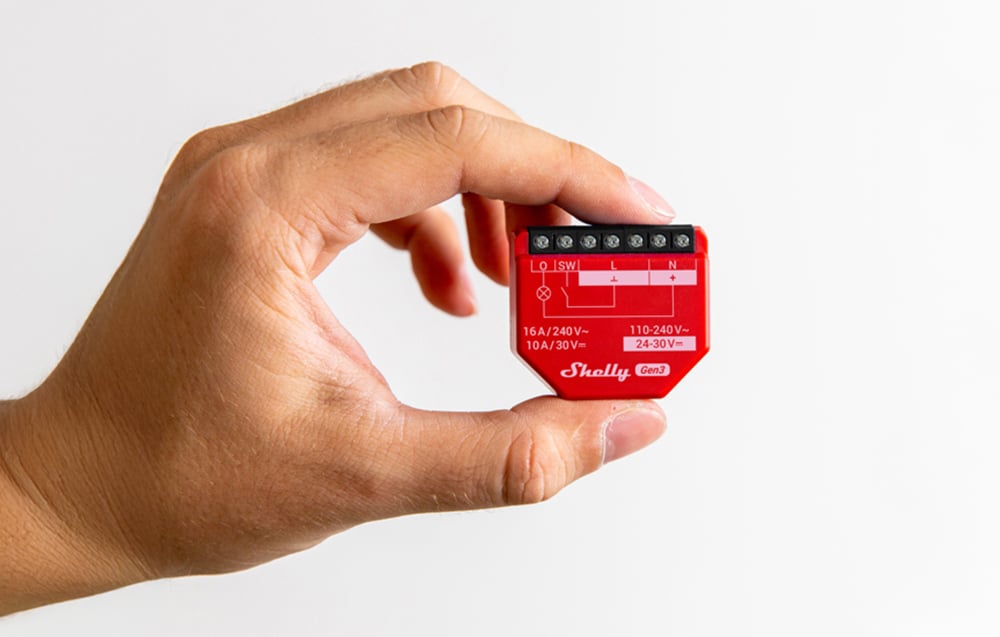 A hand holding a red Shelly 1PM Gen3 module against a white wall background