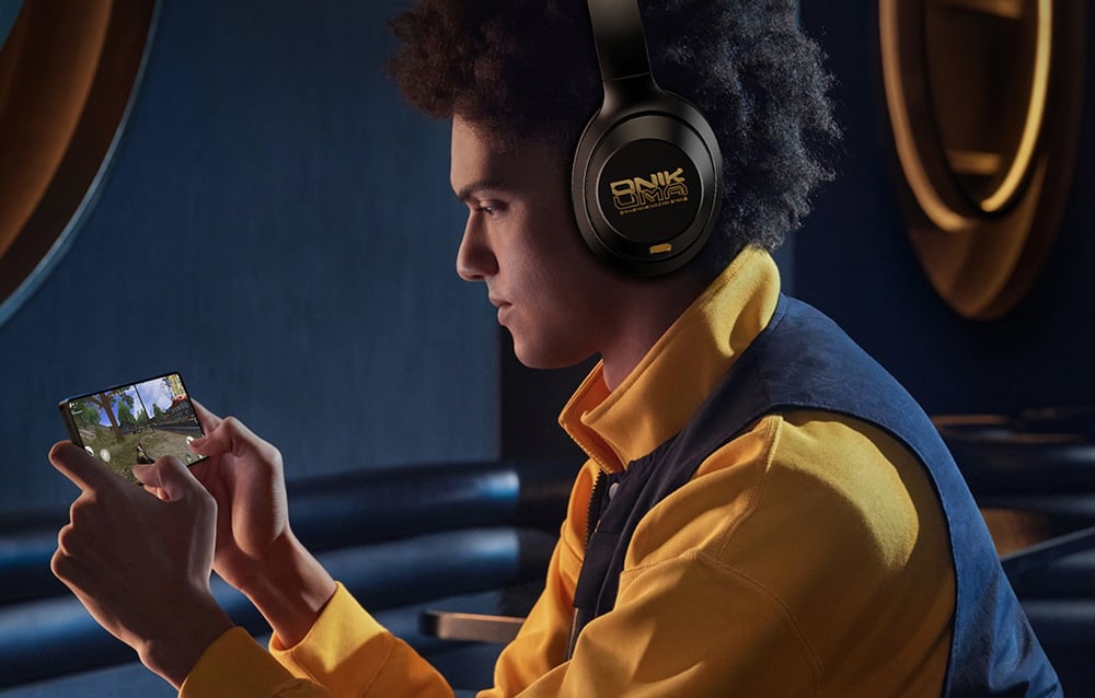 A man in a yellow and blue sweatshirt playing a mobile game and wearing Onikuma headphones