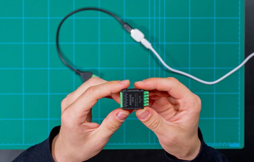 Hands holding the device with “The Pill” written on it above a green cutting mat next to cables