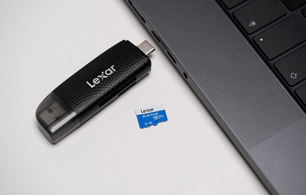 Lexar Blue Plus microSDXC memory card lying on a white tabletop next to a black USB-C card reader and the edge of a gray laptop.