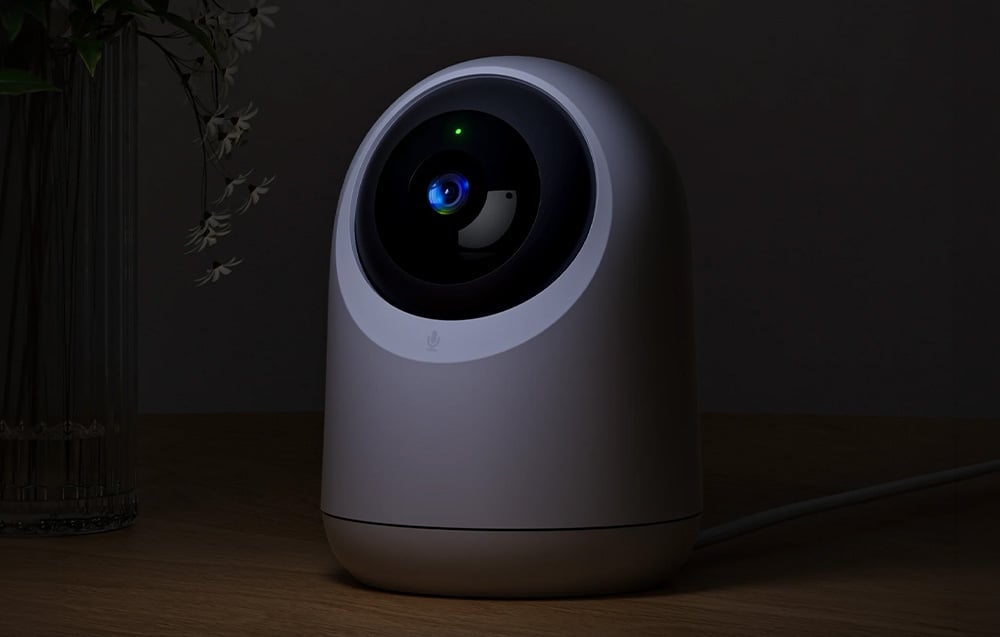 A round white security camera standing on a wooden table in a dark room with a visible green light.