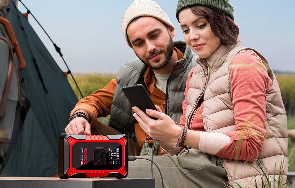 A couple wearing hats is sitting by a tent and charging a phone from a red Gooloo power bank