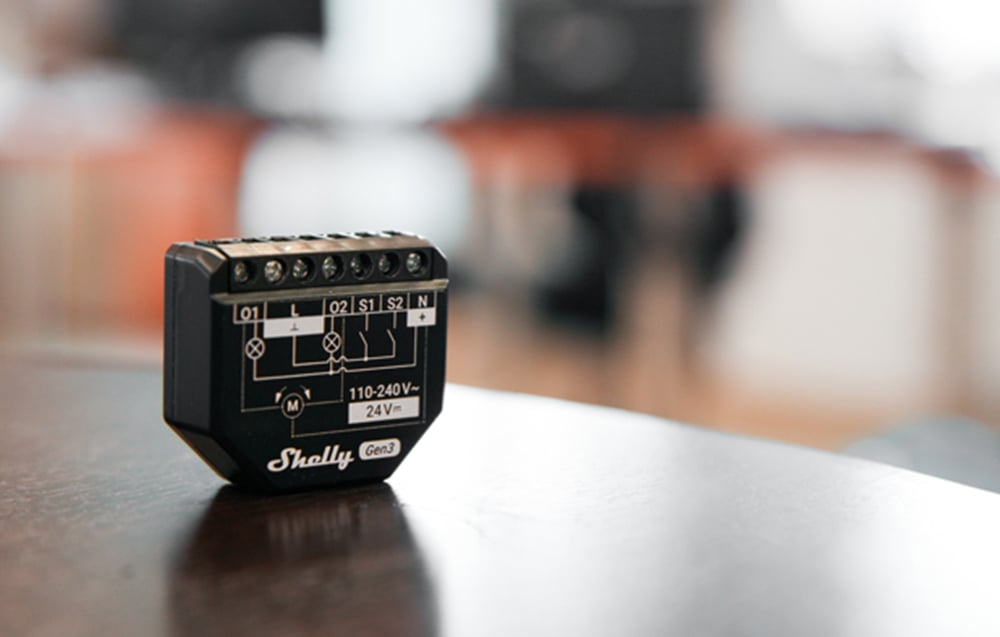 A black Shelly 2PM Gen3 module stands on a dark table, with visible connection markings and power specifications