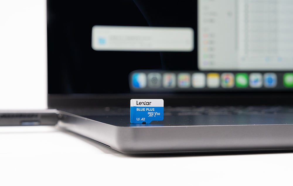 Close-up of the Lexar Blue Plus microSD card standing upright on a laptop case, with a blurred screen displaying the system interface visible in the background.