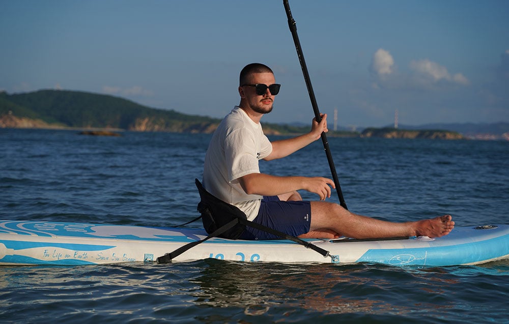 A man wearing sunglasses is sitting on a surfboard with a seat and holding a paddle in his hand