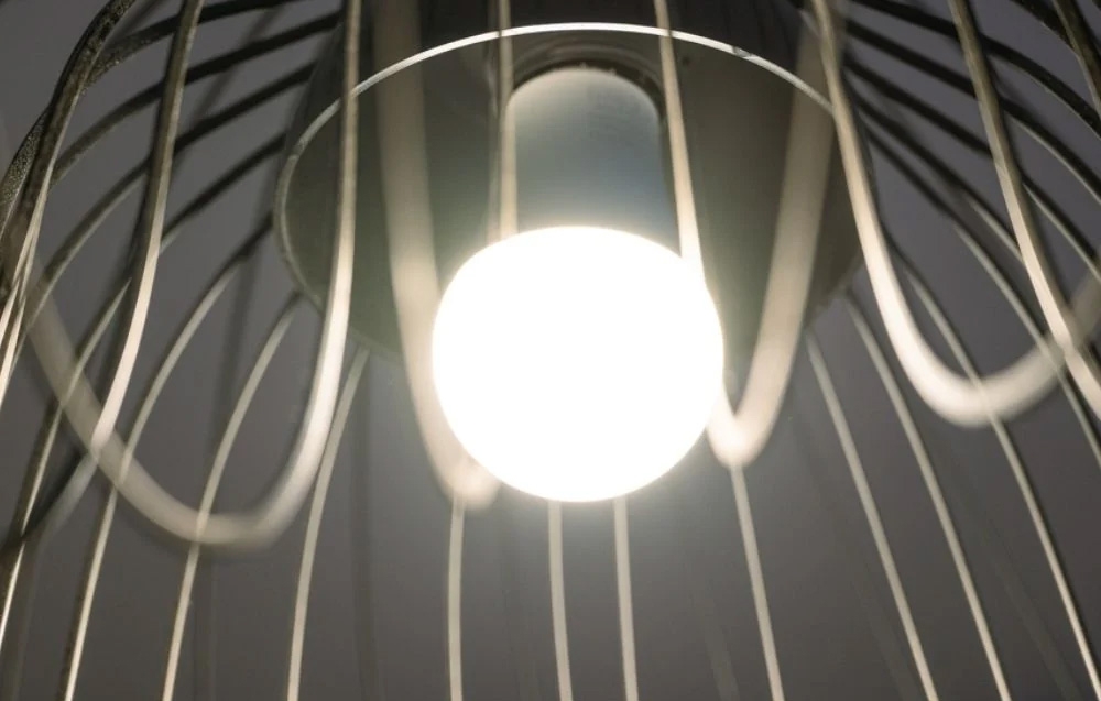 A close-up view from below of a warm light bulb lit through the metal, wire cage of a chandelier