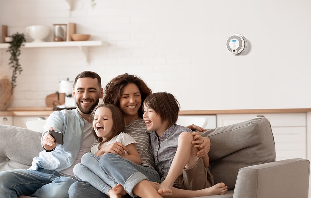 A happy family taking a group selfie on the living room couch, with a white carbon monoxide detector visible on the wall