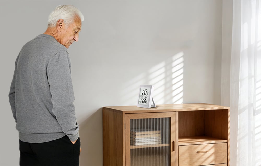 An elderly man looks at the temperature and humidity sensor placed on a chest of drawers in a bright room.
