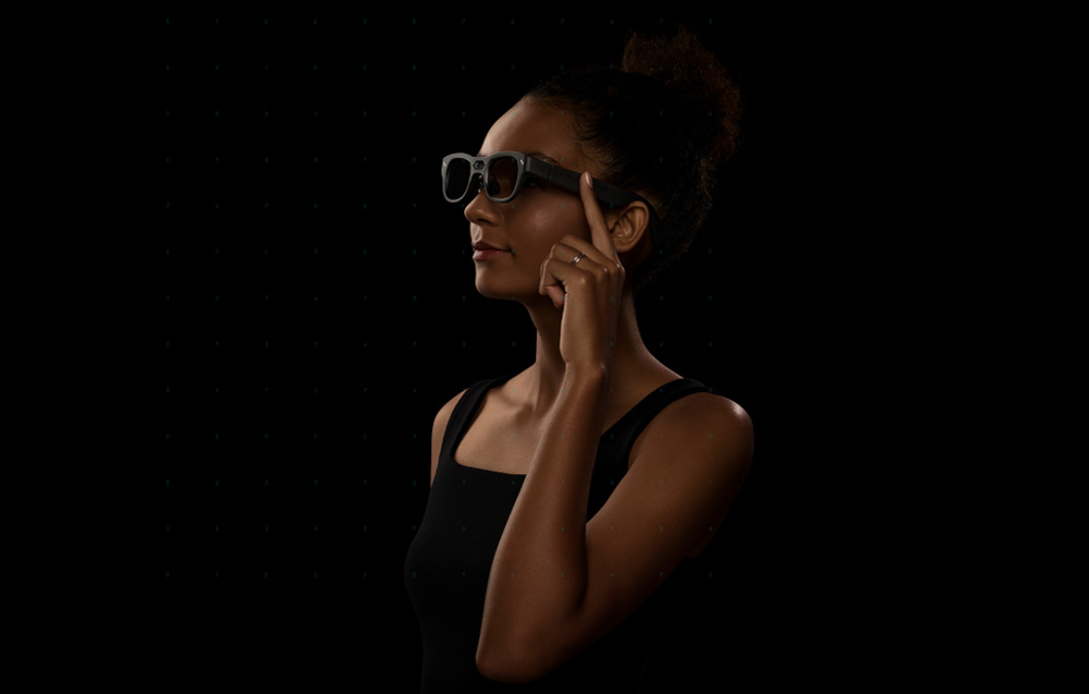 A woman in a black outfit touching the side of the AR glasses against a dark background.