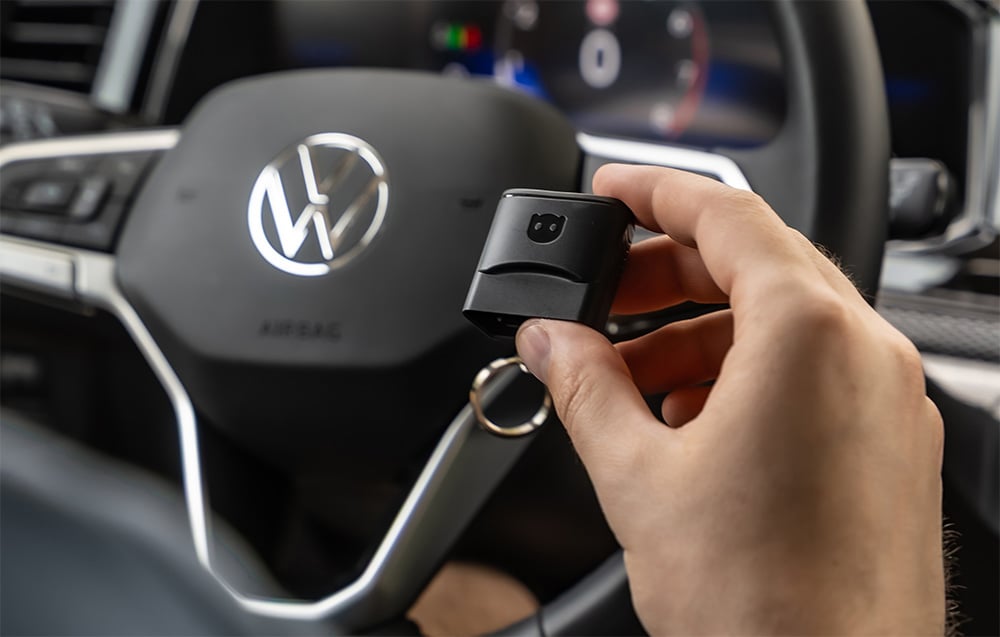 Hand holding Carly adapter in front of Volkswagen steering wheel with VW logo visible