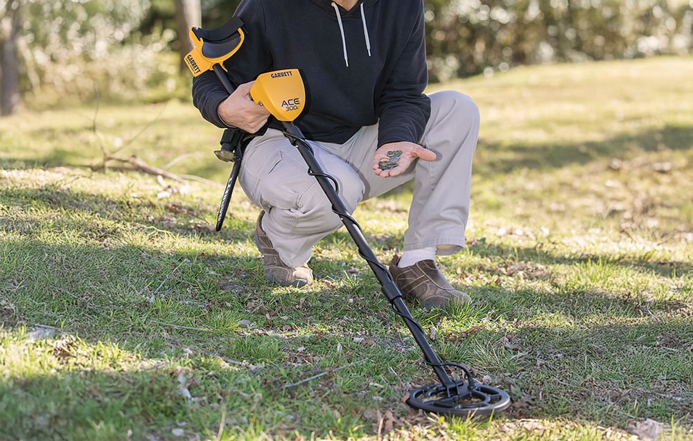 A person kneels on the grass and holds the metal detector and found coins in his hand.