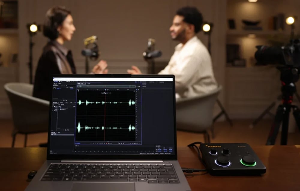 A laptop displaying sound waveforms next to a Maono mixer against the blurred background of a recording studio
